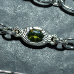 A close-up of a silver chain bracelet featuring a textured, wave-like bezel setting holding a faceted olive green gemstone, displayed on a textured gray background.
