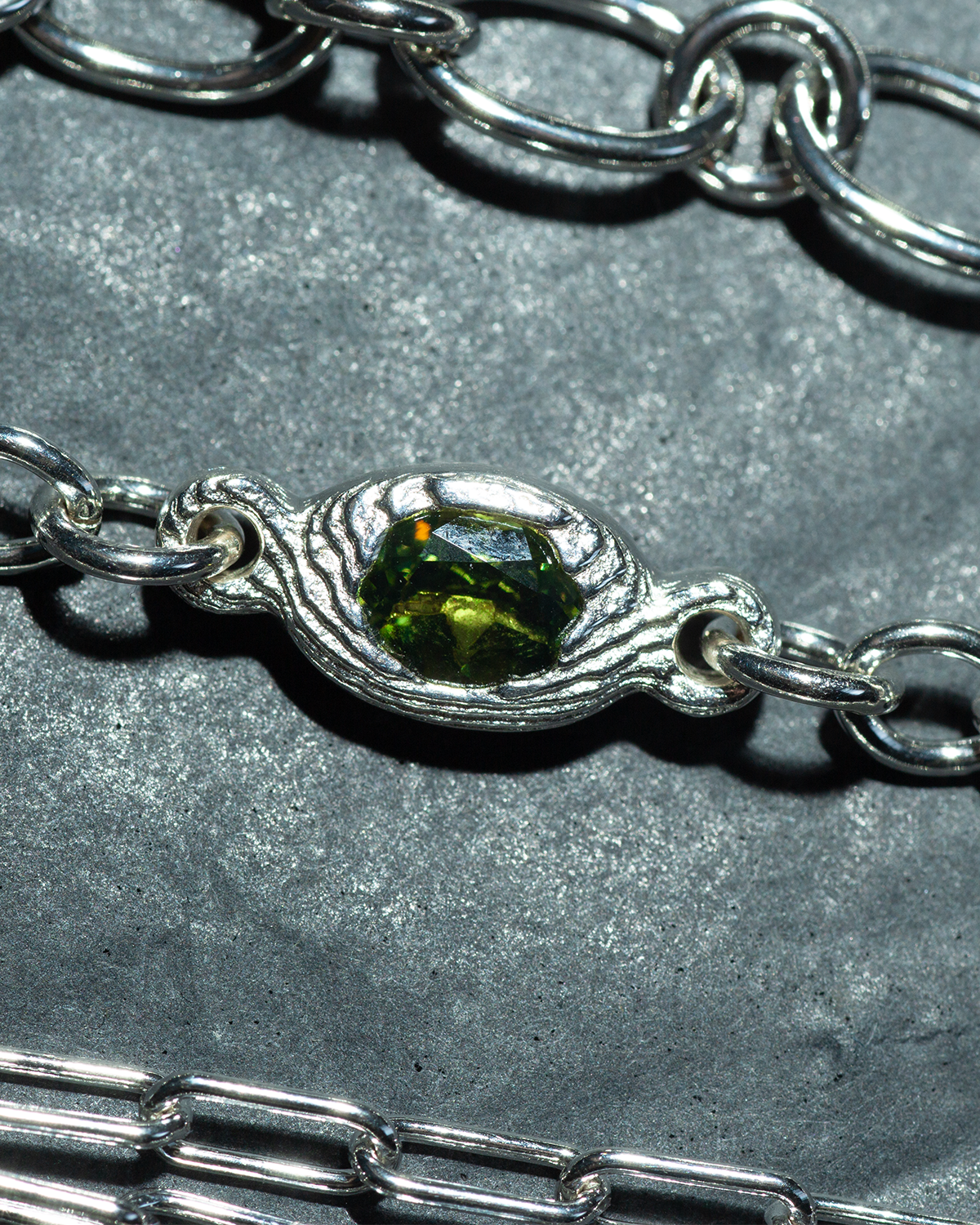 A close-up of a silver chain bracelet featuring a textured, wave-like bezel setting holding a faceted olive green gemstone, displayed on a textured gray background.