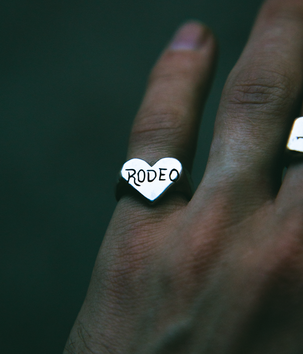 A silver heart-shaped ring engraved with the word 'RODEO' in black lettering, worn on a person's finger against a dark green background.