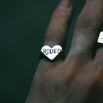 A silver heart-shaped ring engraved with the word 'RODEO' in black lettering, worn on a person's finger against a dark green background.