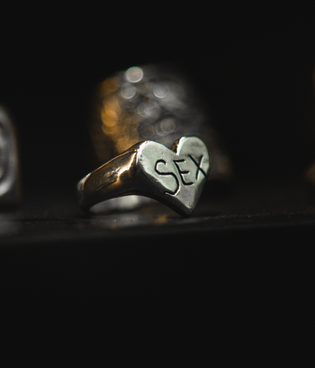 A silver heart-shaped signet ring engraved with the word 'SEX' in black lettering, resting on a dark surface with a blurred gold ring in the background.