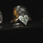A silver heart-shaped signet ring engraved with the word 'SEX' in black lettering, resting on a dark surface with a blurred gold ring in the background.