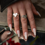 A close-up of a hand with long nude manicured nails wearing two silver rings, each engraved with the word 'MORE' inside a heart shape. The hand is resting near a reflective surface with a dark jacket sleeve visible at the top.
