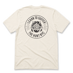 An off-white short-sleeve t-shirt shown from the back, featuring a large black circular graphic print with a cartoon monster face and the text 'LEARN TO SUFFER' and 'THE HUNT NYC'.
