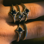 A close-up of a hand wearing two ornate gold rings with dark blue gemstones in a star-like setting.