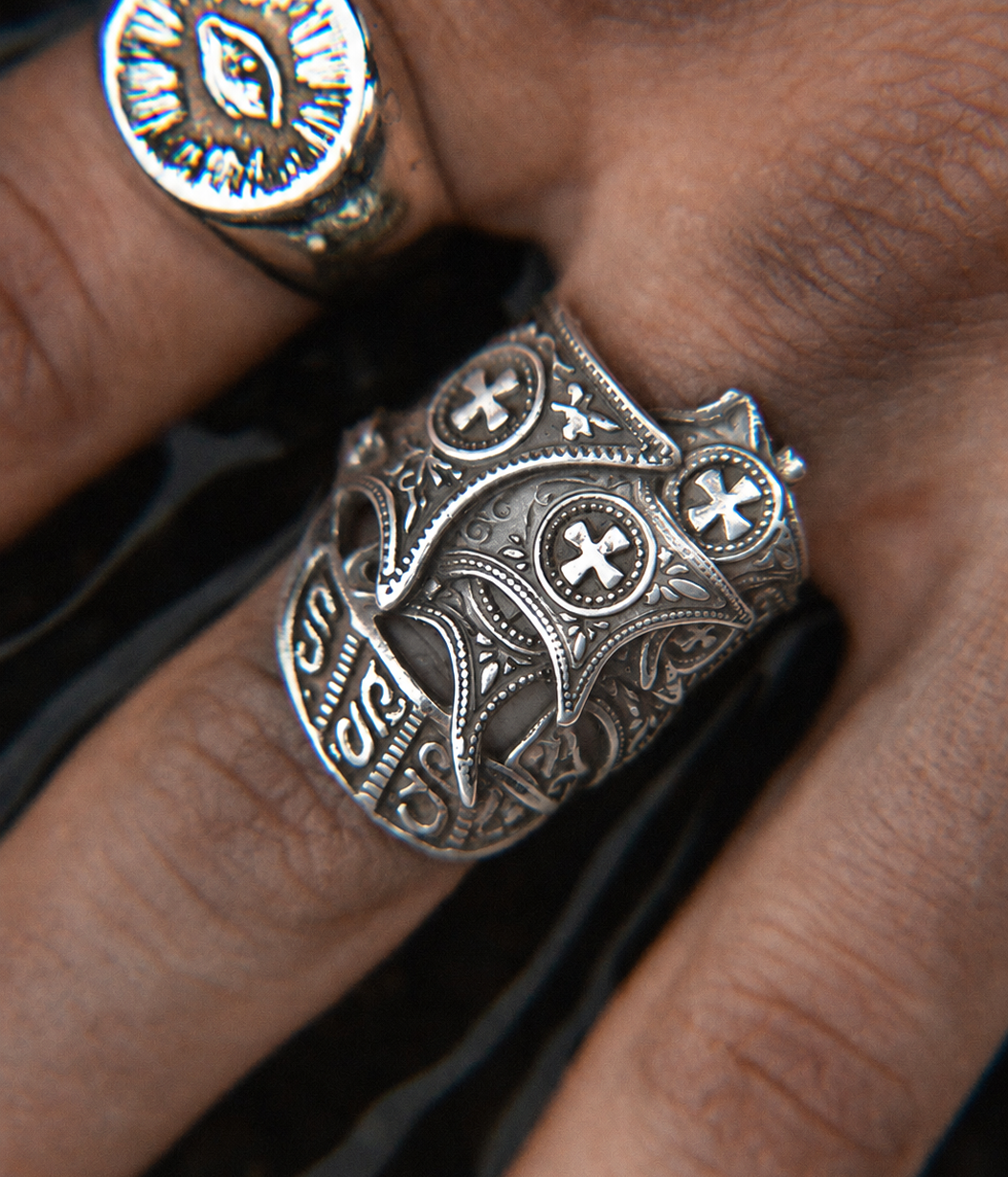 An ornate silver-toned ring with intricate filigree, cross motifs, and engraved details, shown worn on a finger. A second ring is partially visible on another finger in the background.