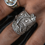 An ornate silver-toned ring with intricate filigree, cross motifs, and engraved details, shown worn on a finger. A second ring is partially visible on another finger in the background.