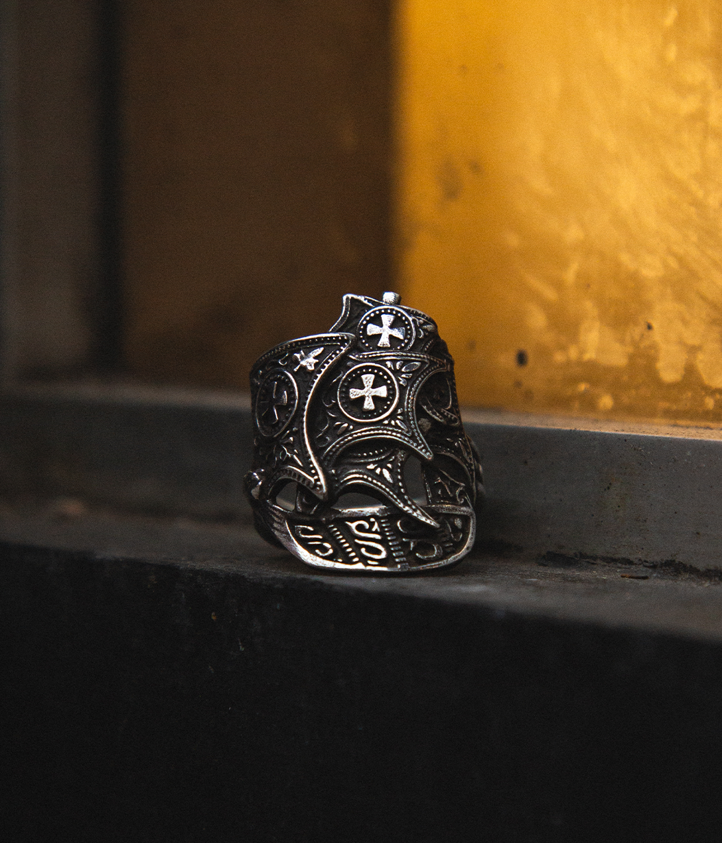 An ornate silver-toned ring with intricate filigree cutouts and cross motifs, resting on a dark ledge against a warm, textured golden-yellow background.