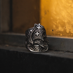 An ornate silver-toned ring with intricate filigree cutouts and cross motifs, resting on a dark ledge against a warm, textured golden-yellow background.