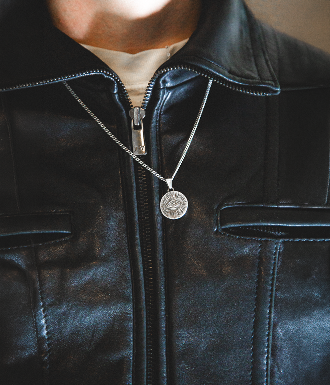 A silver coin pendant necklace with an engraved design hangs around the neck of a person wearing a black leather jacket over a beige shirt.