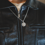 A silver coin pendant necklace with an engraved design hangs around the neck of a person wearing a black leather jacket over a beige shirt.