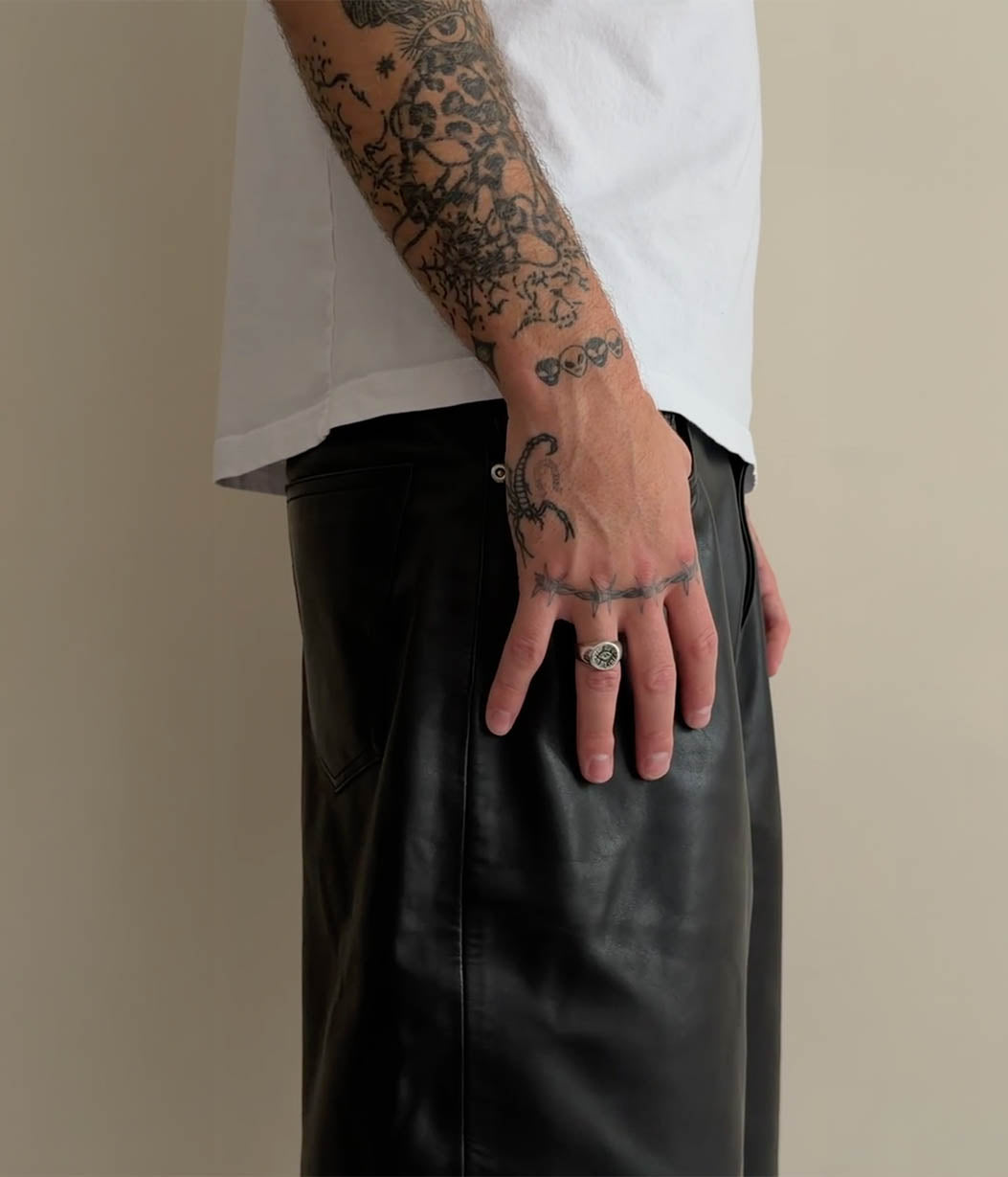 Close-up of a person wearing a white t-shirt and black leather pants, with tattooed arms and hands resting on the hip against a plain beige background.