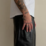 Close-up of a person wearing a white t-shirt and black leather pants, with tattooed arms and hands resting on the hip against a plain beige background.