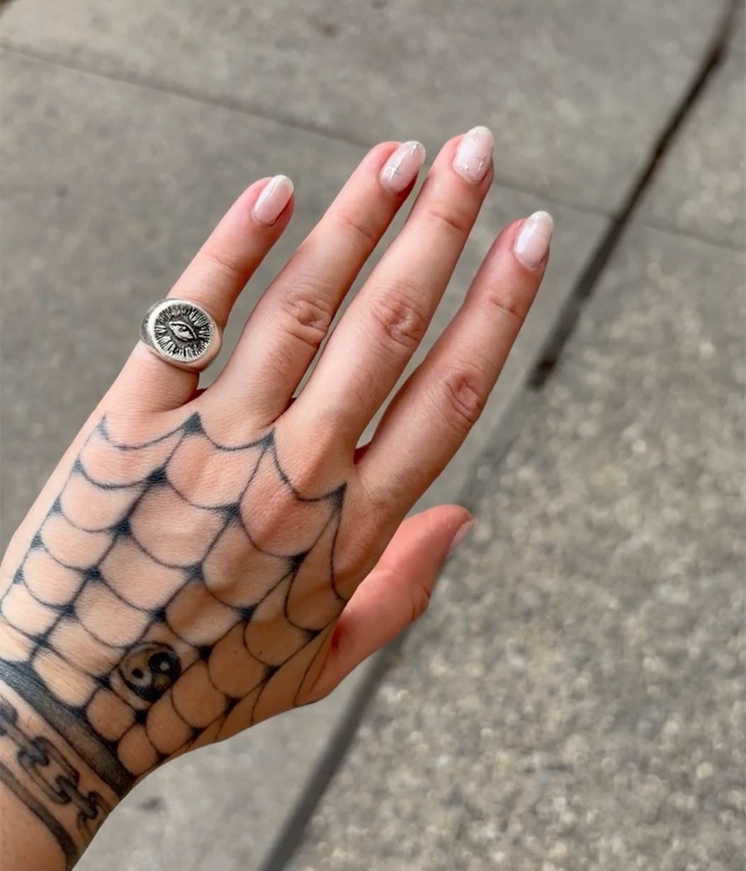 A close-up of a hand with a black spiderweb tattoo and pale pink manicured nails, wearing a silver-toned signet ring with a black skull and crossbones design.