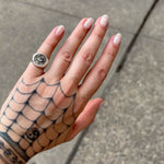 A close-up of a hand with a black spiderweb tattoo and pale pink manicured nails, wearing a silver-toned signet ring with a black skull and crossbones design.