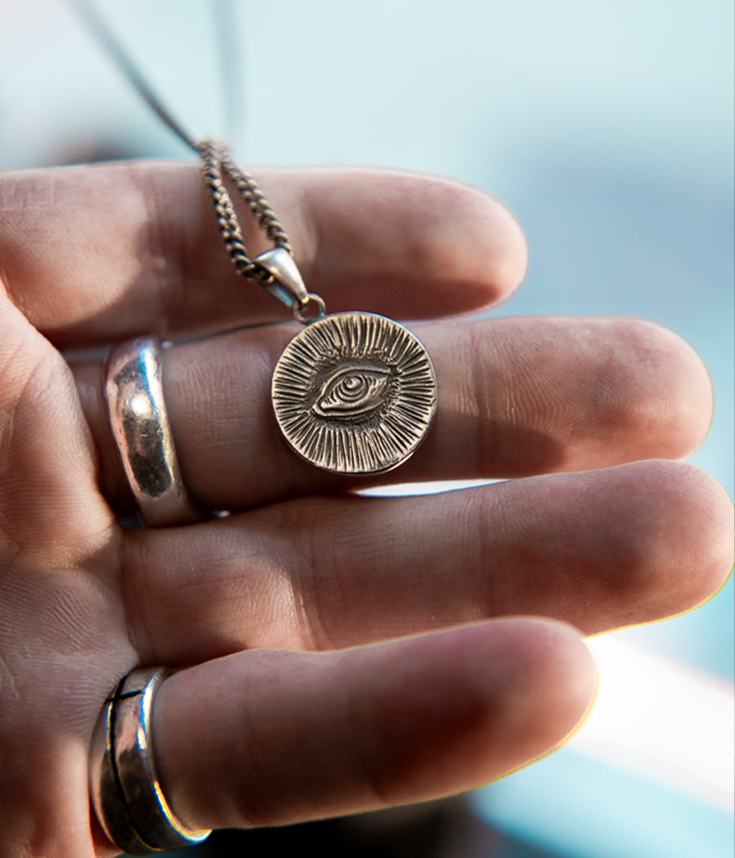 A silver-toned circular pendant necklace with an eye motif, held in a person's hand. The pendant is shown close up against a softly blurred light blue background.