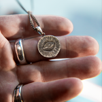 A silver-toned circular pendant necklace with an eye motif, held in a person's hand. The pendant is shown close up against a softly blurred light blue background.